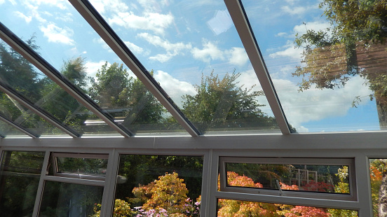 Photo showing a glass conservatory roof with panels of self-cleaning glass.  This glass is slightly tinted and has special properties, helping to reflect the outside heat in the summer and retain the interior heat in the winter.  The flowers, trees and shrubs growing in the summer garden can be seen through the glass roof and windows.