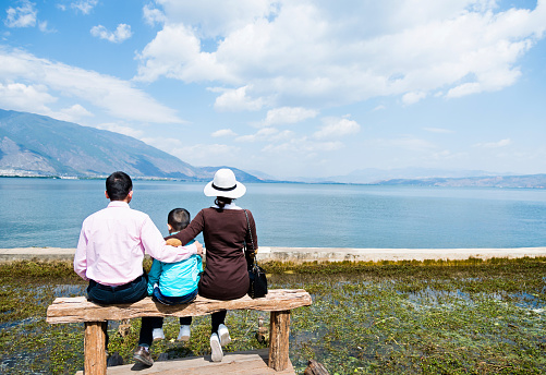 Asian parents and their little son sitting on lakeside jetty.