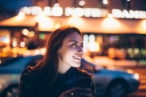 Cheerful young women using smart phone on street by night. Looking away. Street lamps and building lights on background. Focus on foreground.