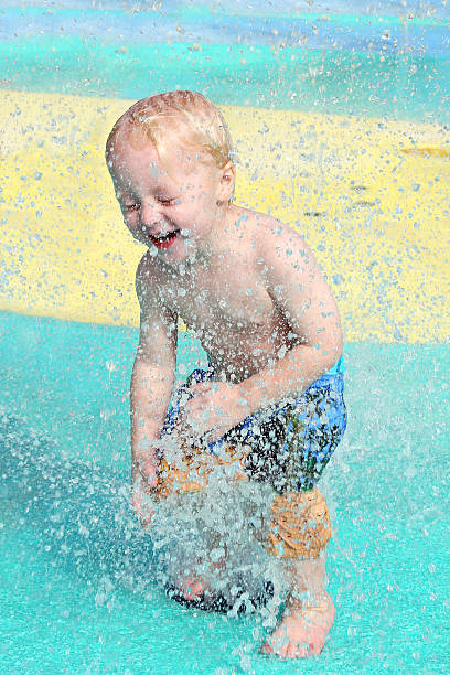A happy young child is laughing as water sprays in his face and gets him wet at a toddler splash pool at a park on a summer day.