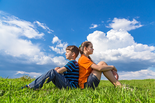 Two kids sitting back-to-back on green grass against blue sky