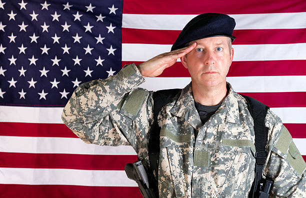 istock Veteran solider saluting with USA flag in background while armed 532879526