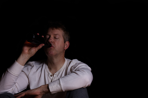 Depressed man drinking beer out of a bottle while sitting down in a dark background.