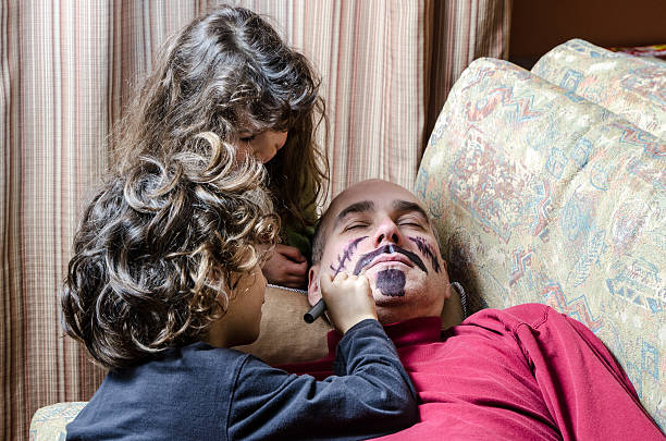 Two kids (boy and girl) are having fun drawing a moustache and a beard on their father's face while he his sleeping on the couch,