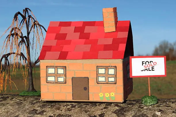 Photo showing a homemade model dolls house made from cardboard and coloured paper.  Standing outside of the front door is a For Sale sign - black writing on a white background, with a red 'Sold' banner, along with a miniature weeping willow tree (salix babylonica).   The toy cardboard house and For Sale / Sold sign are pictured isolated against a rich blue sky.