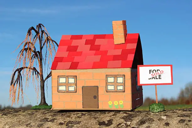 Photo showing a homemade model dolls house made from cardboard and coloured paper.  Standing outside of the front door is a For Sale sign - black writing on a white background, with a red 'Sold' banner, along with a miniature weeping willow tree (salix babylonica).   The toy cardboard house and For Sale / Sold sign are pictured isolated against a rich blue sky.