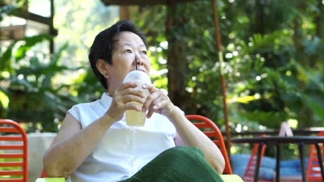 Asian senior woman drinking coffee happily in green lush nature coffee shop