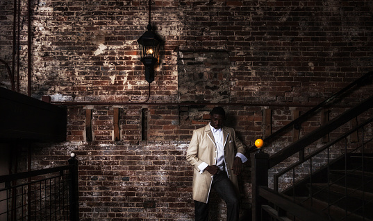 A handsome young black man with his eyes closed is relaxed - resting as he stands waiting for nightlife friends at the bottom of a dark staircase in a rustic old retro revival urban brick wall building. He's wearing contemporary, fashionable, dapper formal clothes - with a long, stylish light-colored tuxedo jacket over a white vest/waistcoat and white dress shirt. Accented with white jacket buttons, a white bow tie and a folded white pocket square handerkerchief. With well-worn, comfortable jeans down below. He's looking fine - ready to head upstairs and join the party.