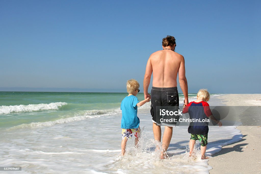 Father and Sons Holding Hands Walking Through Ocean A father and his two young children, both sons, are holding hands as they walk along a white sand beach, through the ocean while on summer vacation. 2015 Stock Photo