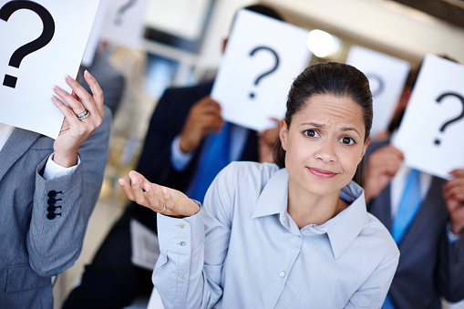 Shot of a group of businesspeople holding up signs with question marks on them during a work presentation while their colleague looks confused