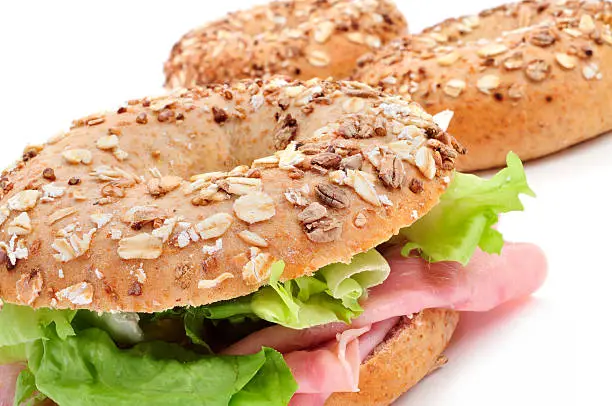 a brown bagel topped with different seeds, such as sesame and poppy seeds, filled with ham and lettuce mix on a white background
