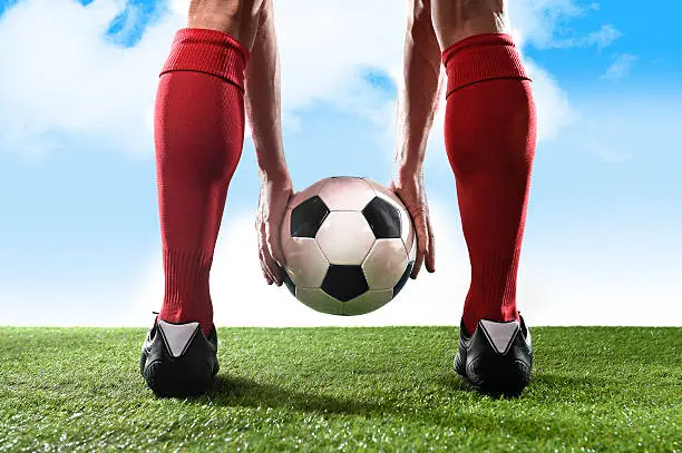 close up legs of football player in red socks and black shoes holding the ball in his hands placing it at the free kick or penalty spot playing outdoors on green grass pitch under a blue sky