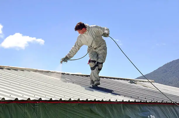 A trademan uses an airless spray to paint the roof of a building