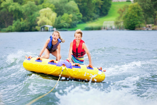 Boy and girl, brother and sister, two active siblings tubing together across a lake in Minnesota, USA. The high speed water sport on an inflatable raft is a family summer vacation activity at Midwest tourist destinations. The children smile playfully as they float across the water surface, flying over waves. 