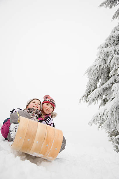 Two happy teen girls/friends sitting on a wood toboggan/sled while at the edge of a snow covered hill in a park.