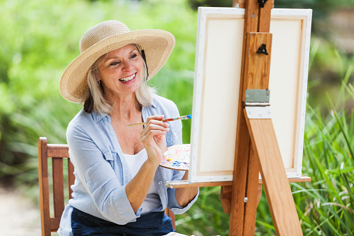 A happy senior woman wearing a wide-brimmed hat is sitting outdoors in a park, in front of a canvas on an easel.  She is holding a palette in one hand and a paint brush in the other.  The background is lush, green foliage.