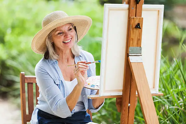 A happy senior woman wearing a wide-brimmed hat is sitting outdoors in a park, in front of a canvas on an easel.  She is holding a palette in one hand and a paint brush in the other.  The background is lush, green foliage.