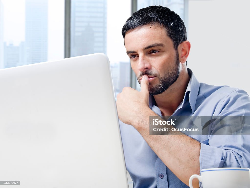 attractive businessman working on computer at office desk Young attractive and handsome businessman working on computer laptop with coffee cup sitting at office desk in front of skyscraper window view looking thoughtful Computer Stock Photo