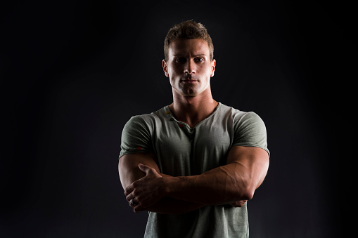 Handsome muscular fit young man on dark background looking at camera, arms crossed on his chest