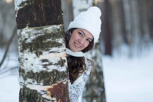 girl in white winter hat hiding behind  tree and smile