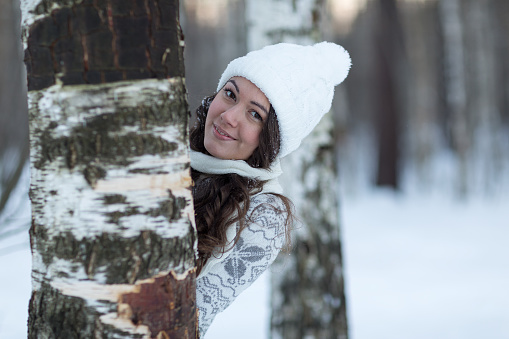 girl in white winter hat hiding behind  tree