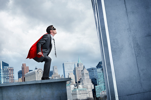 A businessman wearing a cape and a superhero mask about to leap a tall building with a big city skyline in the background. Standing on a cement rooftop, the tall building he faces looms overhead, yet he is not daunted.  With his red cape and black eye mask he stands ready to make the leap. 