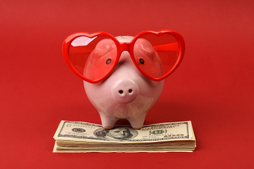 Piggy bank in love with red heart sunglasses standing on stack of money american hundred dollar bills on red background