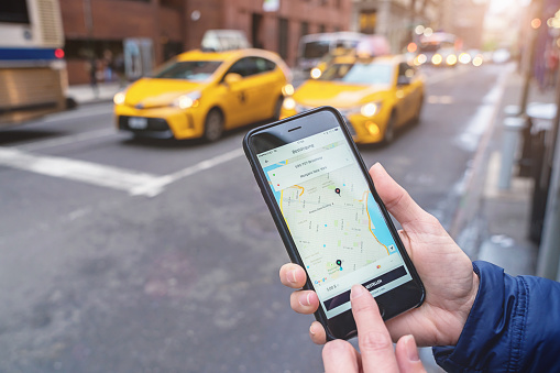 New York, USA - May 6, 2016: Using the Uber taxi App on Apple iPhone 6s in the streets of New York City.Classic NYC yellow cabs in the background. Uber develops, markets and operates the Uber mobile app, which allows consumers with smartphones to submit a trip request which is then routed to Uber drivers who use their own carr as an alternative to the classic taxi service.