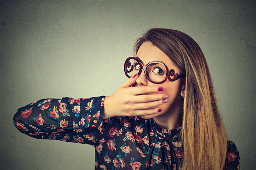 Closeup portrait of scared young woman in glasses covering with hand her mouth isolated on gray wall background. Human face expression emotions feelings .