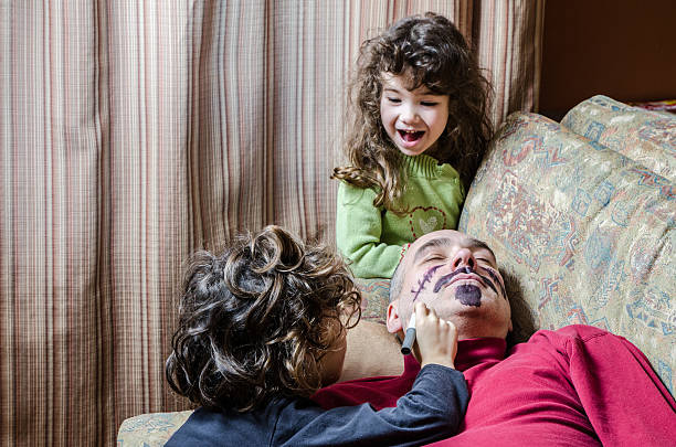 Two kids (boy and girl) are having fun drawing a moustache and a beard on their father's face while he his sleeping on the couch,