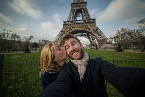 Happy cheerful couple in Paris in front of the Eiffel tower on the Champs de Mars taking a selfie using a mobile phone. Man smiling to camera and woman kissing him on the chick.
