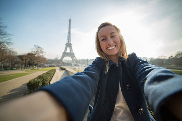 Young cheerful woman near the Eiffel tower  in Paris, France taking a selfie using her mobile phone. Sunny and clear sky day in Winter.