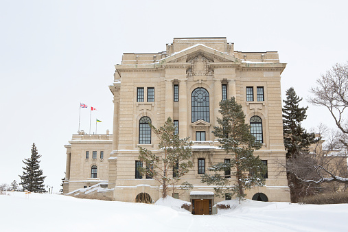 The west entrance of the Saskatchewan Legislative Building in Regina, Saskatchewan in winter. The United Kingdom, Canadian, and Saskatchewan flags fly on the building.