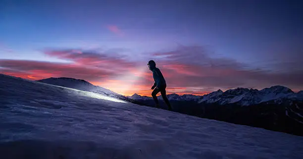 A man walking on the snow with the light of a head lamp, during the sunrise