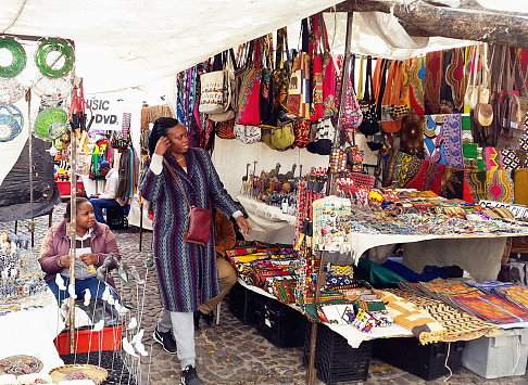 Cape Town, South Africa - May 19, 2016: A shopper walks among stalls selling trinkets and curios, mainly intended for the city's booming tourism trade, in the open-air market in Cape Town's central Greenmarket Square. This daily market features jewellery and curios from many parts of Africa and is a magnet for tourists.