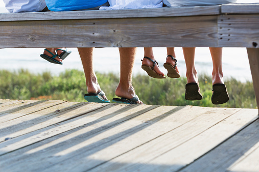 Cropped view of a family with two children sitting on a boardwalk overlooking the beach.  All we see are their legs and feet, wearing sandals and flip flops.