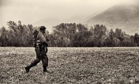 WWII American Soldier On Patrol in a field in Germany.  Rifle ready and moving silently and quickly across an open area.  It is raining and there are mountains in the background.  Copy Space.  Grain and aging of photo.
