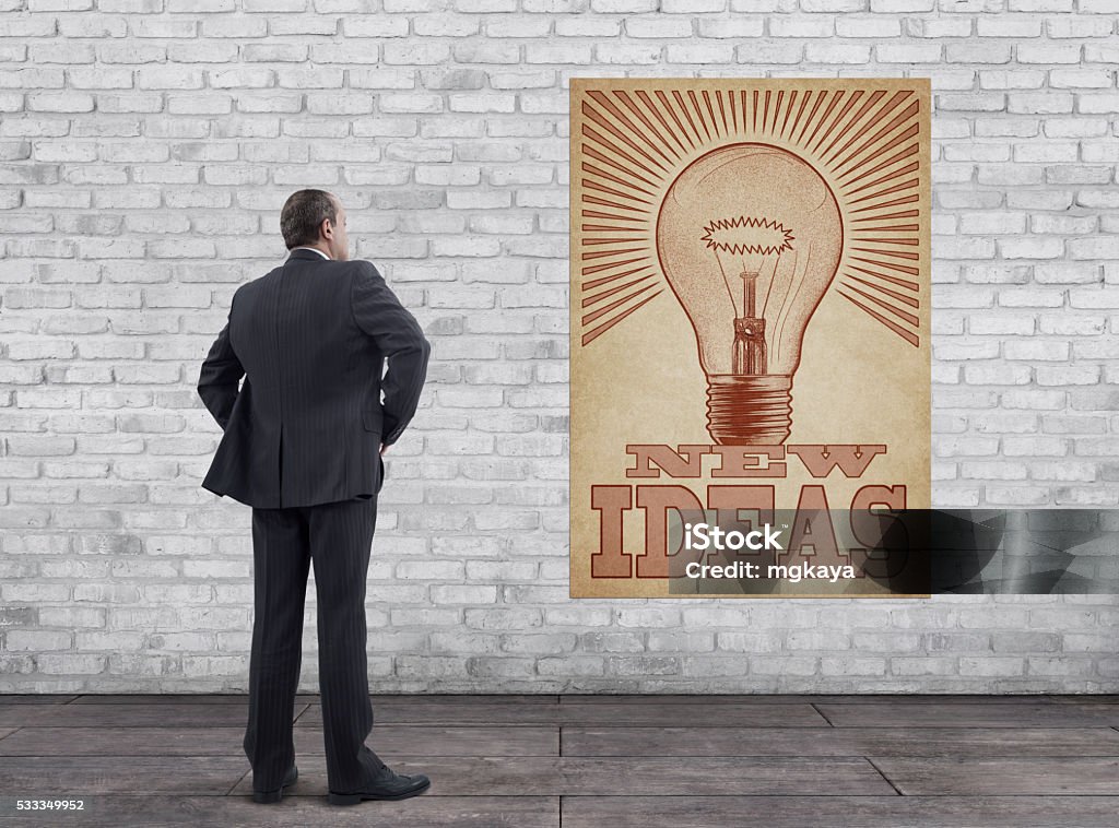 Businessman and “New Ideas” Poster Rear view of businessman standing on wooden floor with vintage style “New Ideas” poster (glowing light bulb illustration printed on old paper) on the white brick wall. Adult Stock Photo