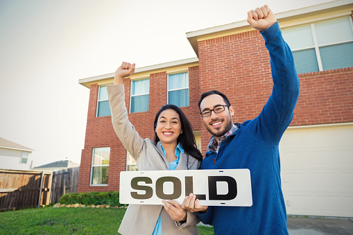 Mid adult Hispanic couple are excited and are cheering with their arms up in the front yard of their new home. They are holding a 'sold' sign. They are standing in front of their two story red brick home with front entry garage. They are dressed in business casual clothing.
