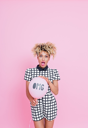 Summer portrait of beautiful afro american young woman with suprised expression on her face. She is wearing grid check playsuit, standing against pink background and holding OMG pink balloon.