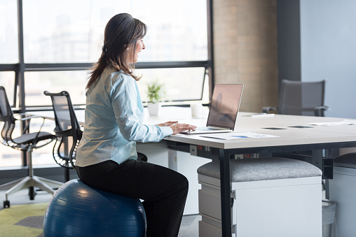 Successful business woman sitting on a Pilates ball and exercising at the office while working