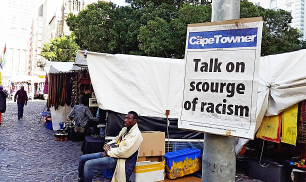 Cape Town newspaper promoting talk on "scourge of racism" Cape Town, South Africa - May 19, 2016:  A stallholder in central Cape Town's open-air market in Greenmarket Square sits near a newsbill for a local free newspaper advertising a talk about racism. More than 22 years after the end of formal racial segregation in South Africa, known as apartheid, the question of racism is firmly back on the political agenda. apartheid sign stock pictures, royalty-free photos & images