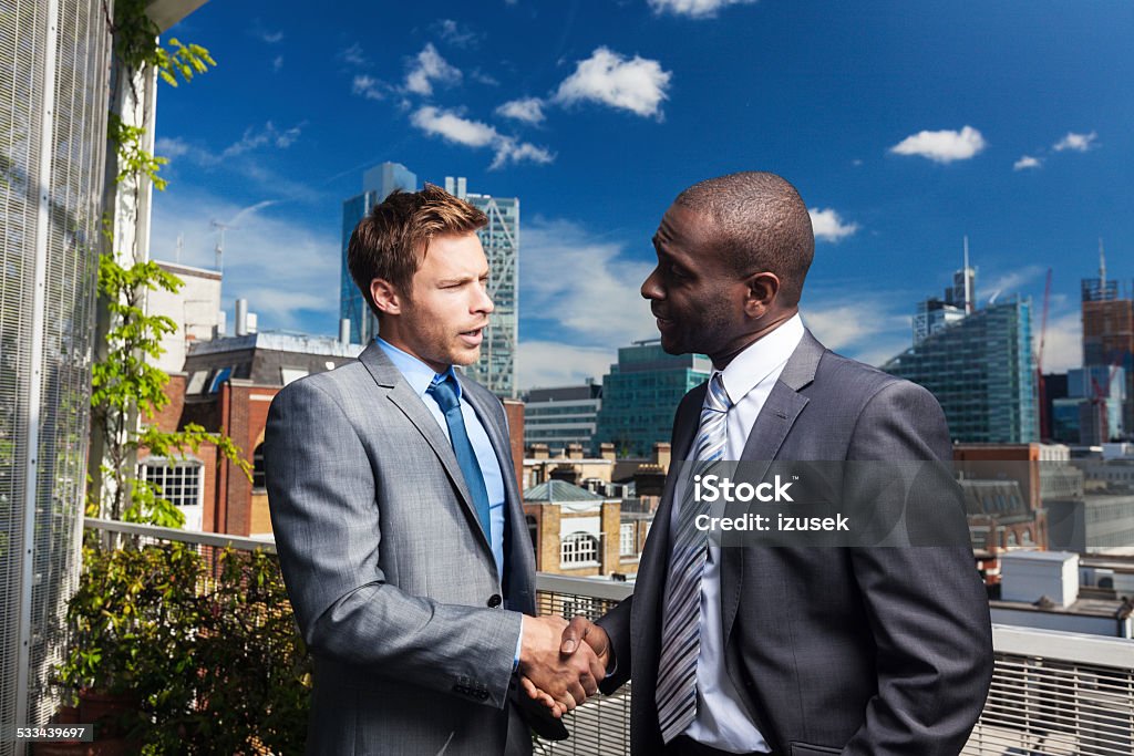 Two businessmen shaking hand outdoors Outdoor portrait of two professional businessmen shaking hands, city scape in the background. African Ethnicity Stock Photo