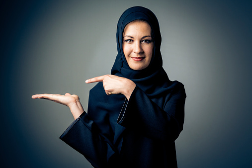 studio shot of young woman wearing traditional arabic clothing. she's holding her hand to the side so you can photoshop in your own content, like a product box or something similar
