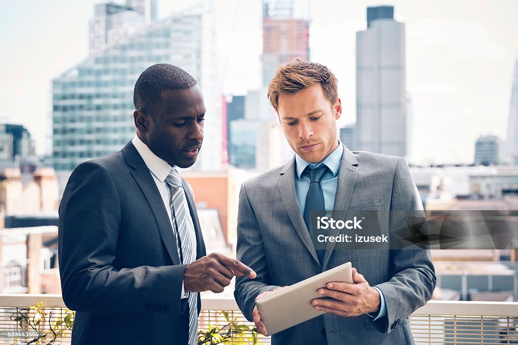 Business people discussing project on digital tablet Afro american businessman discussing project on digital tablet with his business partner, standing outdoor, city scape in the background. Real Estate Stock Photo
