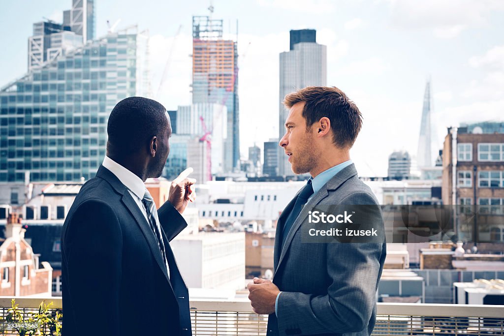 Business people discussing outdoor Afro american businessman discussing with his business partner, standing outdoor, pointing at city scape. Real Estate Stock Photo