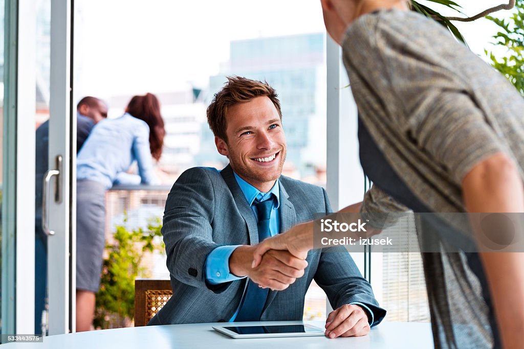 Businessman shaking hands with businesswoman Smiling young businessman sitting in an office and shaking her with his business partner. City scape and business people in the background. Smiling Stock Photo