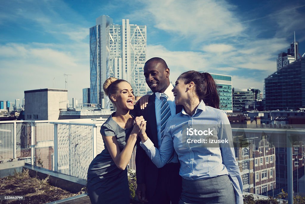 Three happy business people celebrating success Two businesswomen and one businessman standing outdoor on the rooftop of office building and joining their hands, celebrating success. City scape in the background. Corporate Business Stock Photo