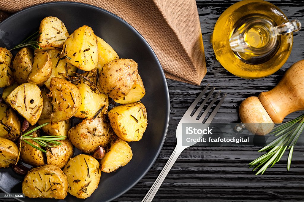 Roasted potatoes Top view of roasted potatoes served in a gray plate on black table. The plate is located at the left of the frame with a fork beside it while a olive oil bottle and a pepper mill are at the top-right. A brown fabric napkin is at the top. DSRL studio photo taken with Canon EOS 5D Mk II and Canon EF 100mm f/2.8L Macro IS USM Roasted Potatoes Stock Photo