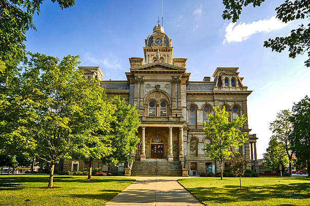 Lavaca County Courthouse with ornate clock tower dominates center of small town square. Historic building, dating from late 19th or early 20th century, surrounded by spacious paved area with brick sidewalks, street lamps, and small trees. Modest commercial buildings line square edges, creating quintessential small-town Texas atmosphere. Scene captures Hallettsville's downtown core, showcasing architectural heritage and community focal point.